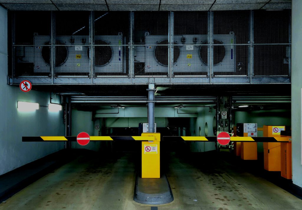 Der Eingang einer Tiefgarage, zwei Schranken, darüber einige Abluftventilatoren. Die Stimmung des Bildes ist durch den beengten Bildausschnitt und den hohen Kontrast dramatisch, eigentlich dramatischer als eine Tiefgarage so ist.
